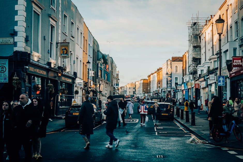 A busy urban street scene during daylight hours with pedestrians crossing and walking along the pavement, surrounded by a variety of shopfronts and buildings. The street is lined with parked cars and 