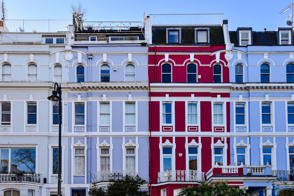 Photograph of a row of Victorian-style terraced houses in Notting Hill with distinctive ornate facades. The buildings feature large windows with white decorative trims and balconies, painted in pastel
