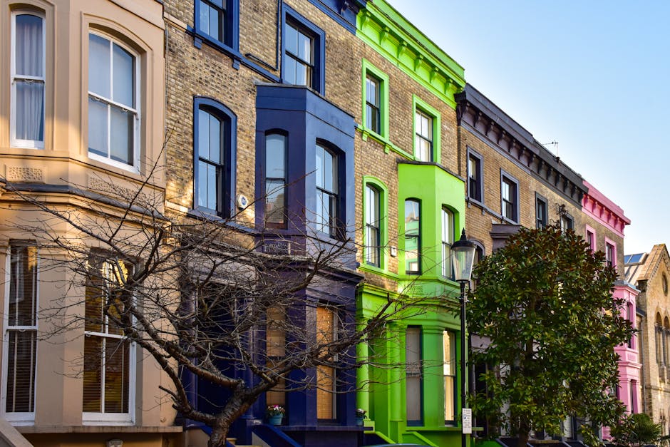 Photograph of a row of Victorian-style terraced houses in Notting Hill with distinctive ornate facades. The buildings feature large windows with white decorative trims and balconies, painted in pastel