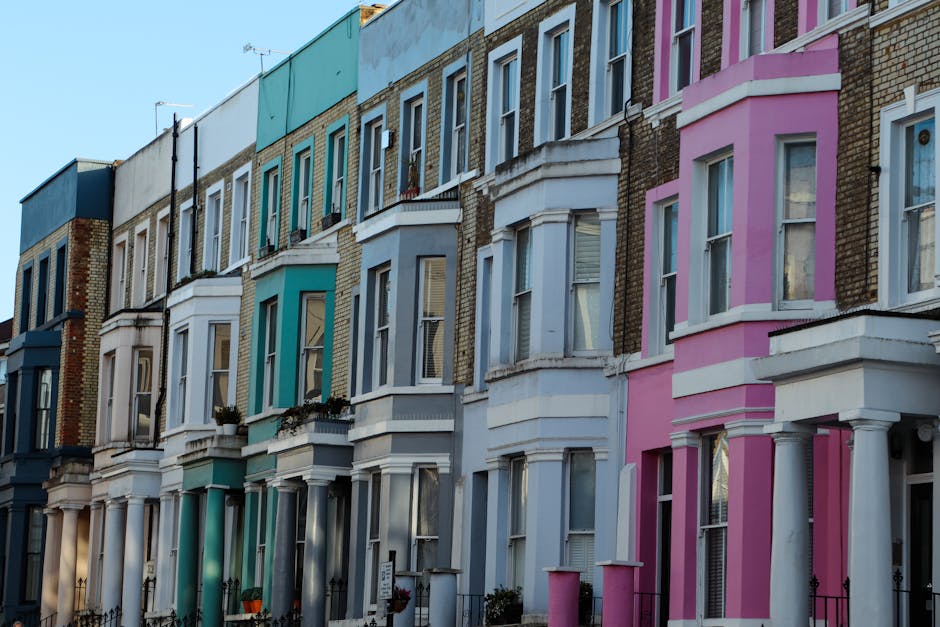 A row of colorful terraced houses on a residential street, each with distinct pastel facade colors including blue, pink, and yellow. The houses feature classic Victorian architectural details such as